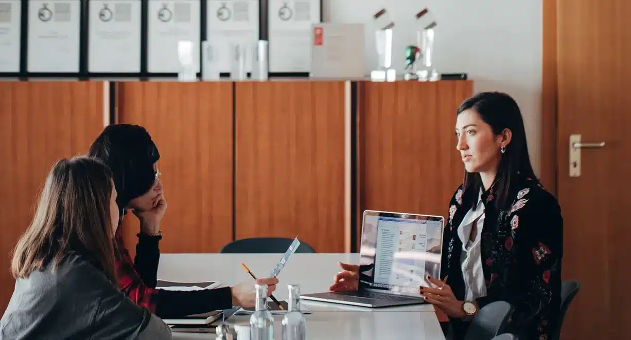 3 Frauen in einem Agentur Meeting an einem Tisch, im Hintergrund ein Schrank mit Awards darauf