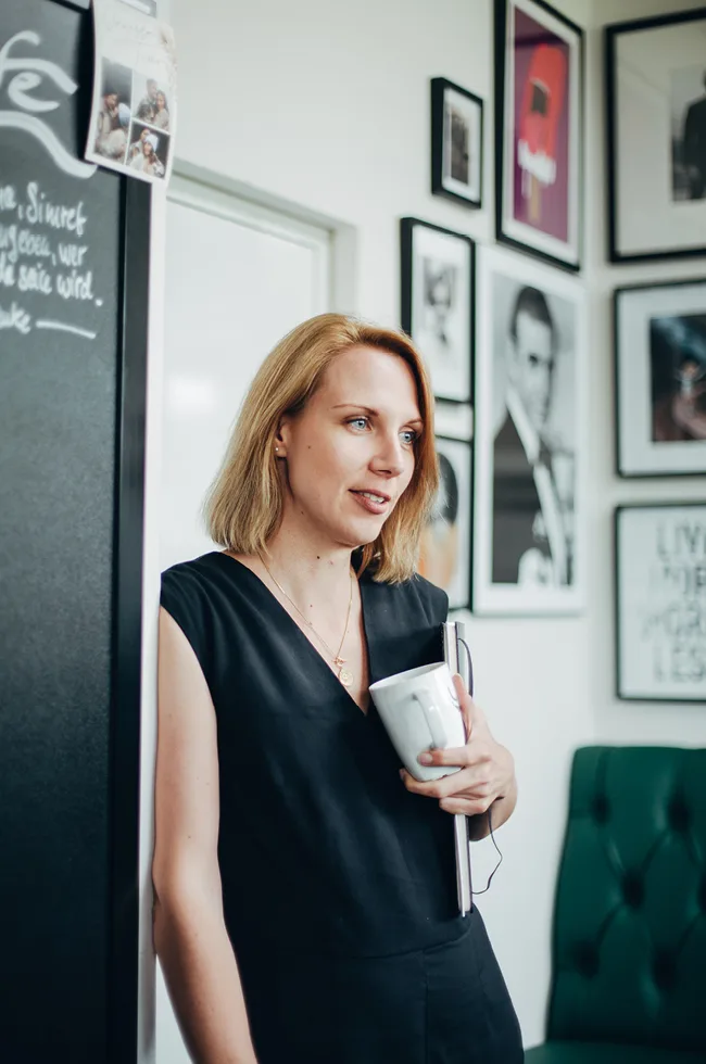 Frau lehnt an einer Wand, Kaffee und Notizbuch in der Hand, Bilder im Hintergrund