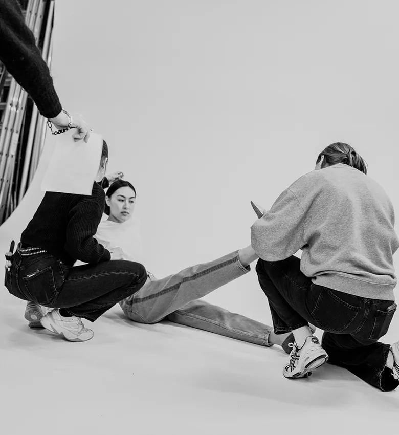 Schwarz-weißes Foto-Shooting für die Marke Reischmann, auf dem ein Model auf dem Boden sitzt und zwei Mitarbeiterinnen es für die Fotos positionieren und frisieren