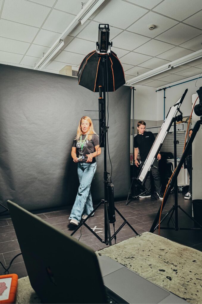 Fotostudio-Setup mit großer Softbox, schwarzem Hintergrund und reflektierenden Lichtpaneelen. Eine Frau mit Mikrofon bewegt sich durch das Set, während ein junger Mann im Hintergrund Lichttechnik anpasst.
