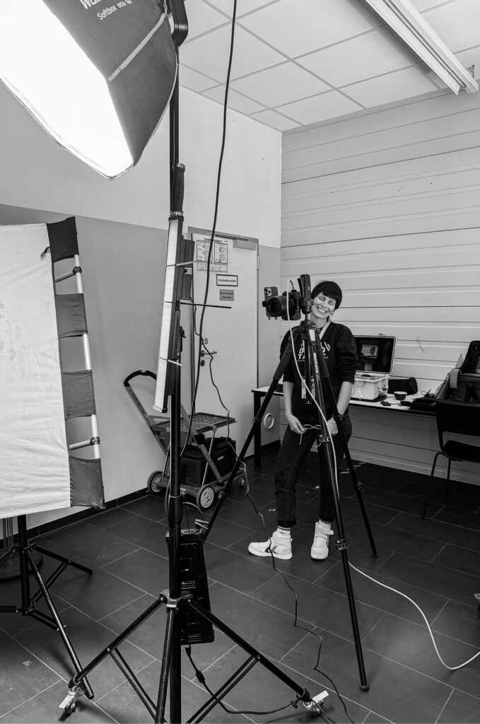 Fotografin mit Kurzhaarschnitt lächelt hinter der Kamera in einem professionellen Fotostudio mit Licht-Setup und Stativen. Schwarz-weiß.