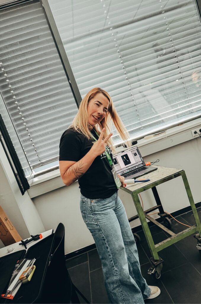 Frau mit blondem Haar und schwarzem Shirt zeigt Victory-Zeichen vor Laptop in Werkstattumgebung. Der Arbeitsplatz in einem Industrie- oder Werkstattumfeld zeigt Werkzeuge und einen Metalltisch mit Rollen.