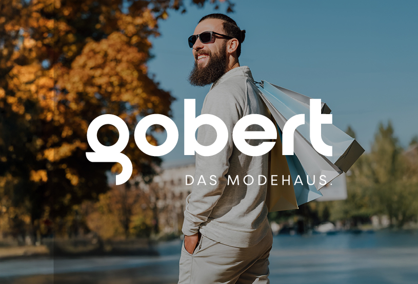 Mann mit Bart, Sonnenbrille und Einkaufstaschen vor herbstlicher Kulisse, darüber das Logo von gobert – Das Modehaus. Beispiel für ein konsistentes Corporate Design mit moderner Typografie, klarer Markenästhetik und starker Bildsprache.
