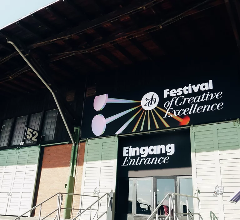 Ein Gebäudeeingang mit einem Schild, auf dem „Festival of Creative Excellence“ und „Eingang“ über einer Doppeltür steht. An der Fassade des Gebäudes sind große Fenster zu sehen, und die Zahl 52 ist erkennbar.