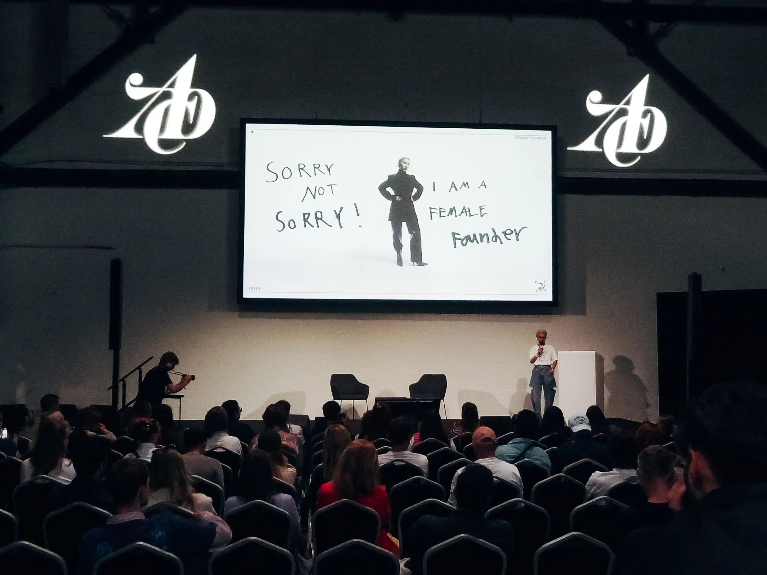 Auf der Bühne steht eine Rednerin vor dem Publikum, hinter ihr ist auf einer großen Leinwand der Text „SORRY NOT SORRY! I am a FEMALE Founder“ zu sehen sowie das Bild einer Frau, die die Hände in die Hüften gestemmt hat.