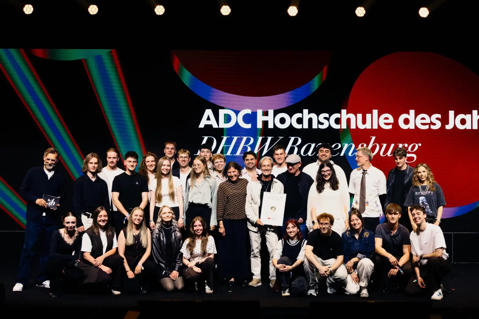 Eine große Gruppe lächelnder junger Erwachsener posiert gemeinsam auf der Bühne, einige halten ein gerahmtes Zertifikat in den Händen. Hinter ihnen ist auf einer bunten Leinwand die Aufschrift „ADC Hochschule des Jahres DHBW Ravensburg“ zu sehen.