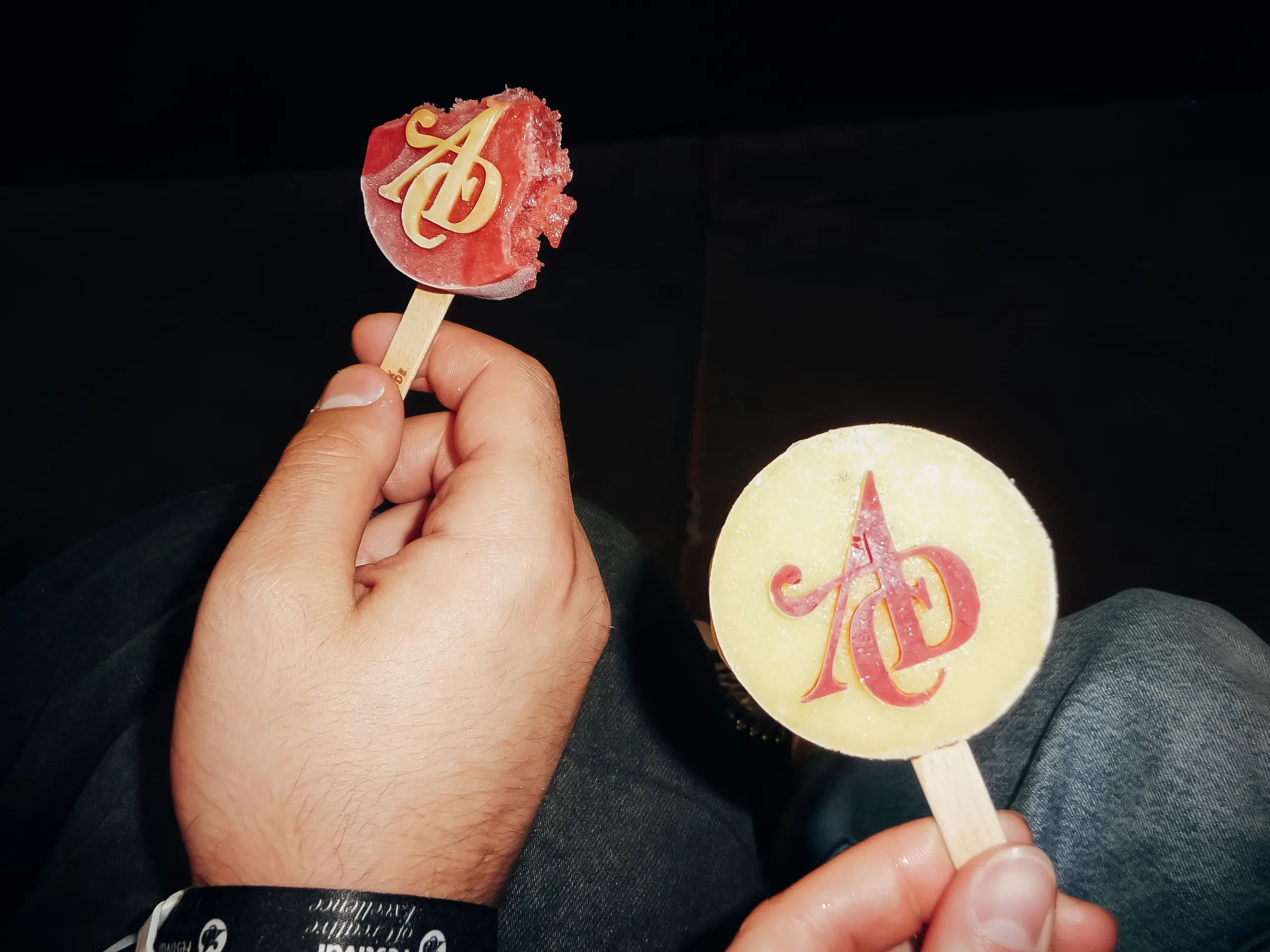 Zwei Hände halten runde Lutscher am Stiel, die jeweils mit einem stilisierten roten AD-Logo versehen sind. Ein Lutscher ist rot und hat einen Bissabdruck, der andere ist gelb und unversehrt. Der Hintergrund ist dunkel.