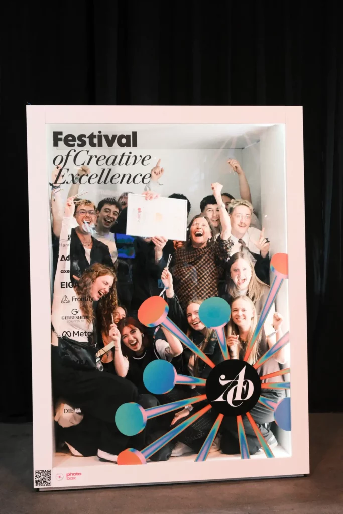 Eine Gruppe von Menschen jubelt und posiert in einem großen Requisiten-Bilderrahmen mit der Aufschrift „Festival of Creative Excellence“, während sie eine Urkunde in den Händen hält und damit feiert.