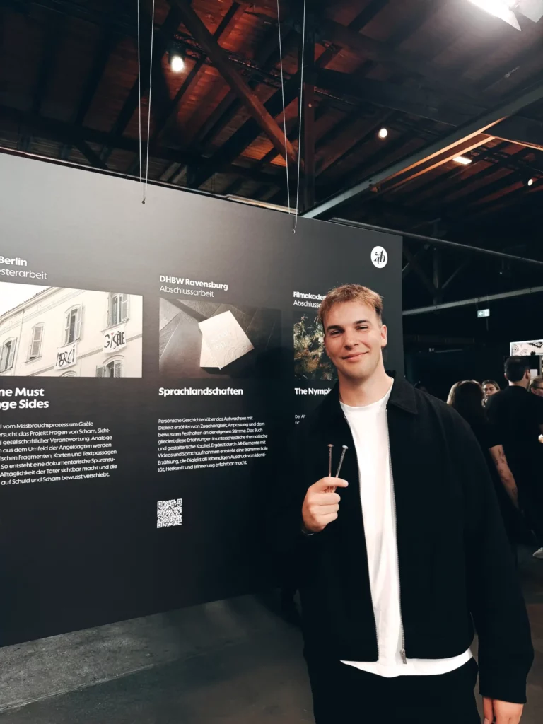 Ein junger Mann in schwarzer Jacke und weißem Hemd steht in einem Raum, lächelt und hält eine Stimmgabel vor einer Ausstellungswand mit Informationstafeln und Bildern. Im Hintergrund sind weitere Personen zu sehen.