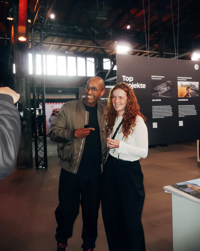 Ein Mann in einer braunen Jacke und eine Frau in einem weißen Langarmshirt posieren gemeinsam und lächeln für ein Foto bei einer Veranstaltung in einem Innenraum, im Hintergrund sind Informationsschilder und eine helle Beleuchtung zu sehen.