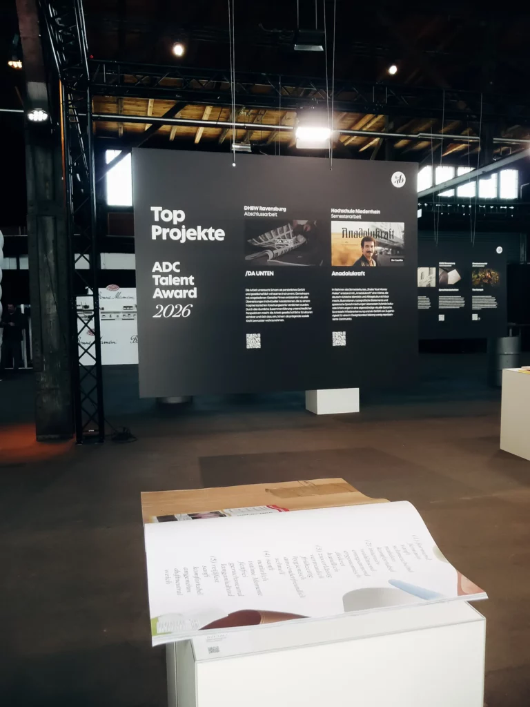 Auf einer großen schwarzen Ausstellungstafel steht „Top-Projekte ADC Talent Award 2026“, darunter befinden sich Projektbeschreibungen. Im Vordergrund liegt ein aufgeschlagenes Buch auf einem weißen Sockel. Im Hintergrund ist ein industrieller Innenraum zu sehen.
