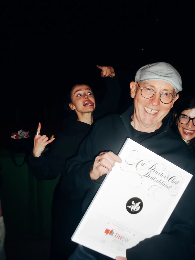 Ein lächelnder Mann mit Brille und grauer Mütze hält eine Urkunde in der Hand, während zwei Frauen hinter ihm spielerisch posieren: Die eine zeigt nach oben, die andere macht ein „Rock“-Handzeichen. Der Hintergrund ist dunkel.