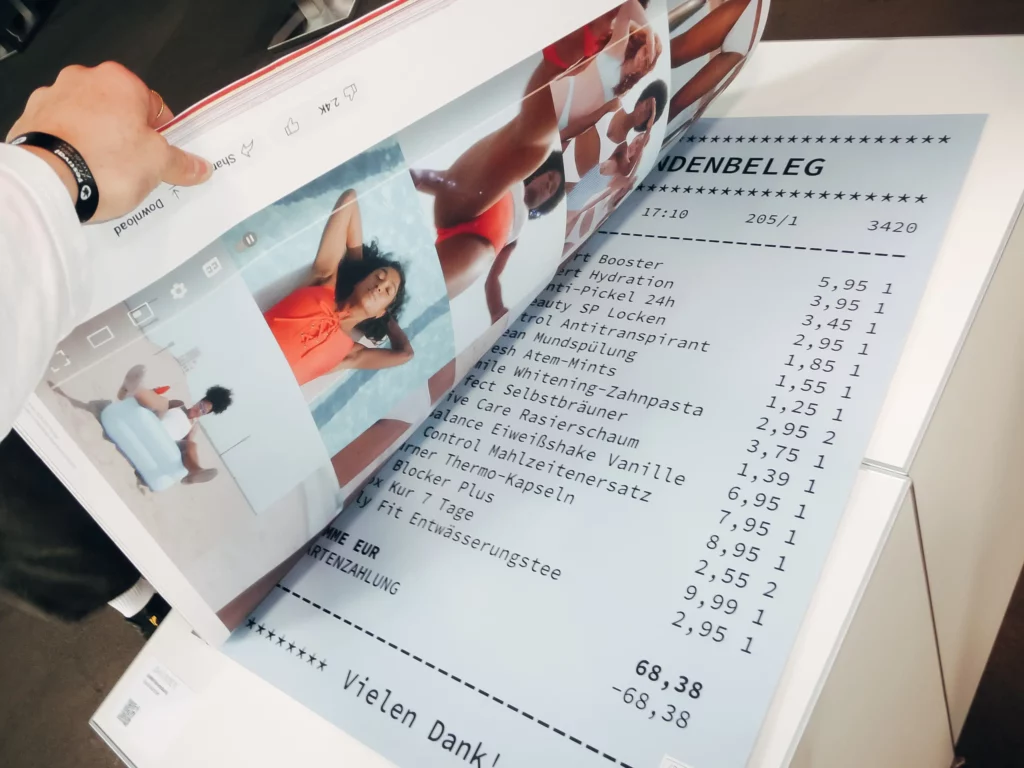 Eine Person blättert in einem großen Buch mit Bildern von Frauen in Badeanzügen und enthüllt dabei einen überdimensionalen, ausgedruckten Supermarkt-Kassenbon, der unter den Seiten hervorschaut.
