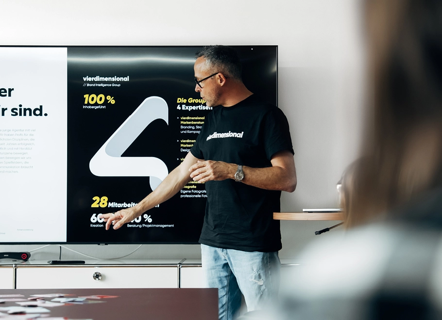 Ein Mann mit Brille und schwarzem T-Shirt zeigt auf eine große Präsentationsleinwand, auf der Statistiken und Informationen auf Deutsch angezeigt werden, wobei Zahlen und Grafiken besonders hervorgehoben sind – sie verdeutlichen die wichtigsten Punkte einer Checkliste für den Relaunch einer Website.