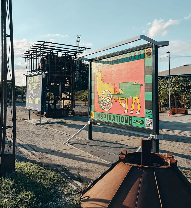 Kunstausstellung im Freien mit einem farbenfrohen Plakat, das ein Pferd und einen Wagen zeigt und mit der Aufschrift „INSPIRATION“ versehen ist. Daneben steht ein Schild mit der Aufschrift „WELCOME. CELEBRATE DIVERSITY & CREATIVITY“. Unter dem blauen Himmel sind Industrieanlagen und Grünflächen zu sehen.