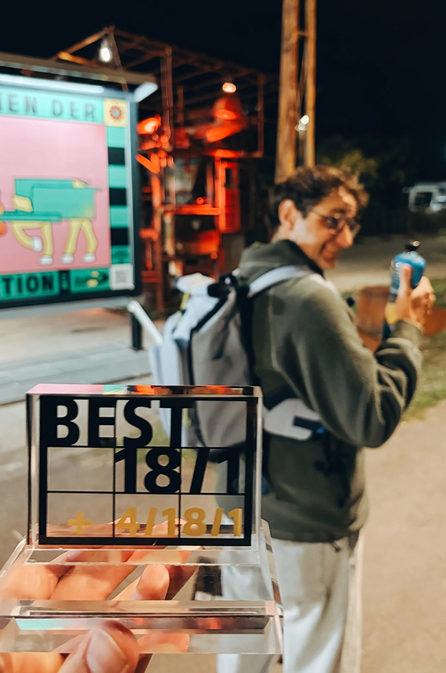 Im Vordergrund hält eine Hand eine „Best 1871 + 4/18/41“-Trophäe in der Hand; im Hintergrund dreht sich eine lächelnde Person mit Brille und Rucksack um und blickt zurück, während sie bei einer nächtlichen Veranstaltung im Freien eine Flasche in der Hand hält.