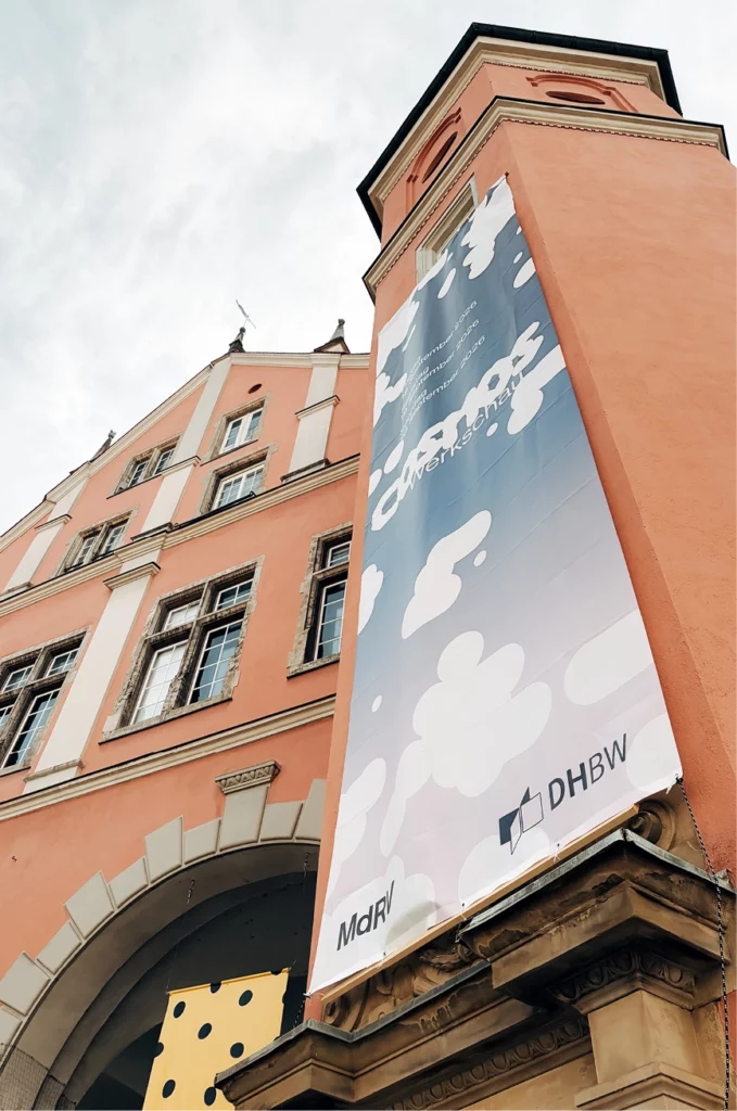 Ein hohes, lachsfarbenes Gebäude mit Bogenfenstern und einem großen, vertikalen Banner mit Wolken, Logos und den Schriftzügen „MdRV“ und „DHBW“ in der Nähe des Eingangs. Der Himmel darüber ist teilweise bewölkt.