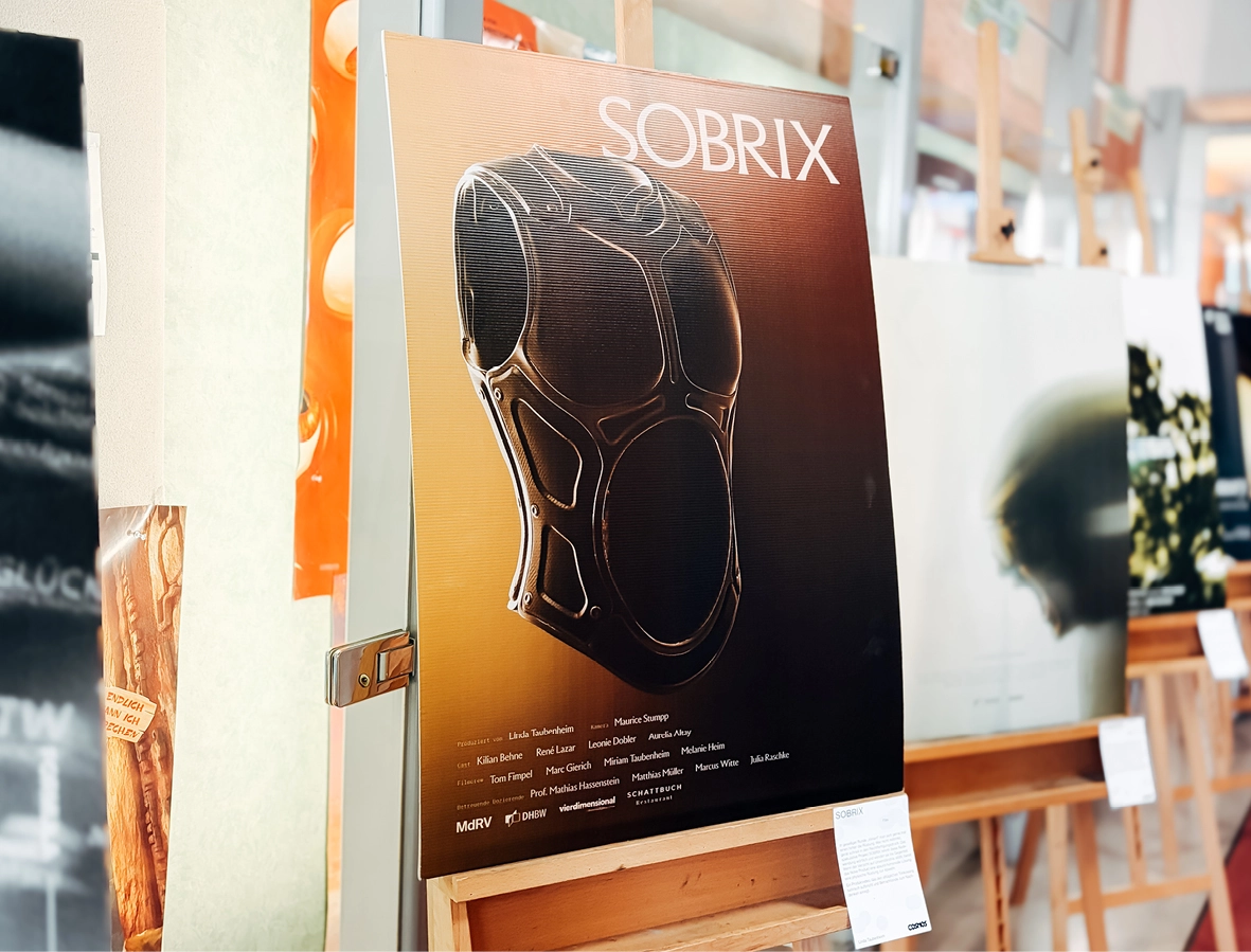 Ein Plakat mit dem Titel „SOBRIX“, auf dem ein futuristisches, schwarzes, brustpanzerähnliches Objekt auf einer Staffelei zu sehen ist, darunter der Filmabspann. Im Hintergrund sind weitere Plakate zu sehen.