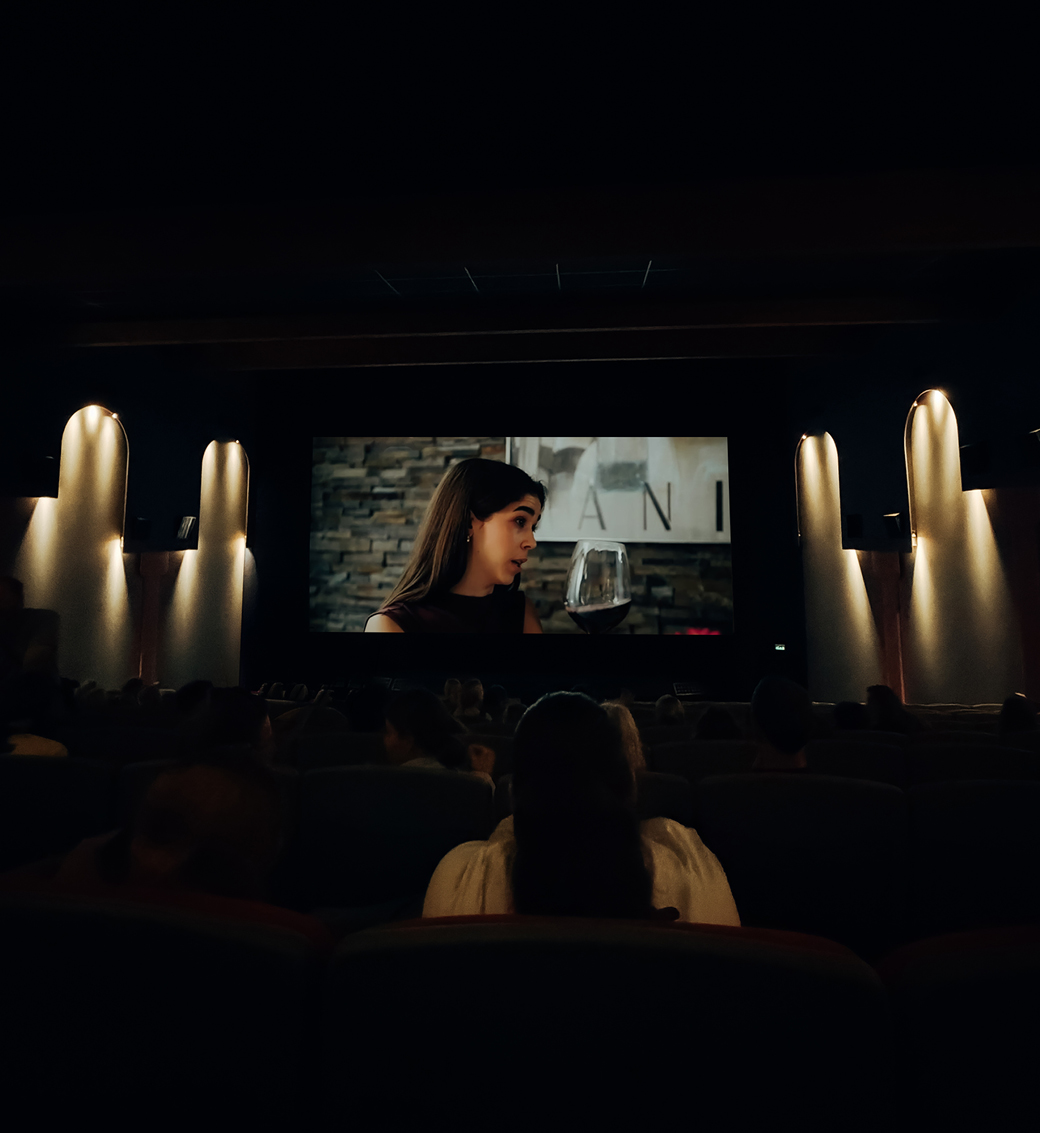 Ein schwach beleuchtetes Kino, in dem Menschen sitzen und einen Film auf der großen Leinwand verfolgen, auf der eine Frau zu sehen ist, die spricht und ein Glas in der Hand hält, während im Hintergrund eine Steinmauer zu erkennen ist.
