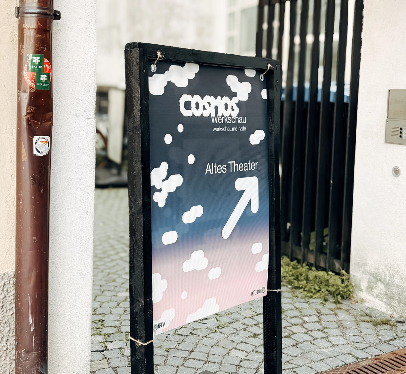 Auf einem Kopfsteinpflaster-Gehweg neben einem Zaun und einer Gebäudewand steht ein Schild mit der Aufschrift „COSMOS Werkschau Altes Theater“ und einem nach rechts zeigenden Pfeil. Das Schild hat einen Hintergrund mit Wolken und einem Farbverlauf.