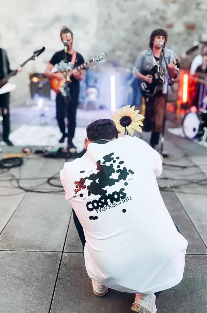 Eine Person in einem weißen Hemd, in dessen Kragen eine Sonnenblume steckt, hockt sich hin und fotografiert eine Band, die im Freien mit Gitarren und Mikrofonen auftritt.