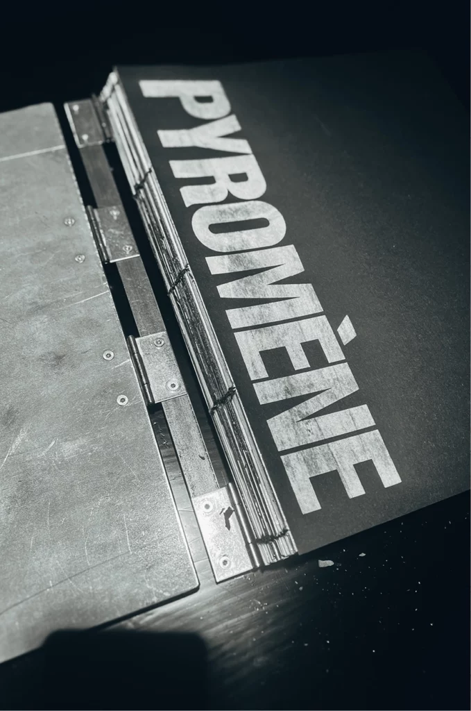 Ein großer Metallordner oder eine Presse hält ein schwarzes Blatt Papier, auf dem das Wort „PYROMÈNE“ in fetten, weißen Großbuchstaben steht. Die Szene ist dramatisch beleuchtet, sodass Schatten auf die Oberfläche fallen.