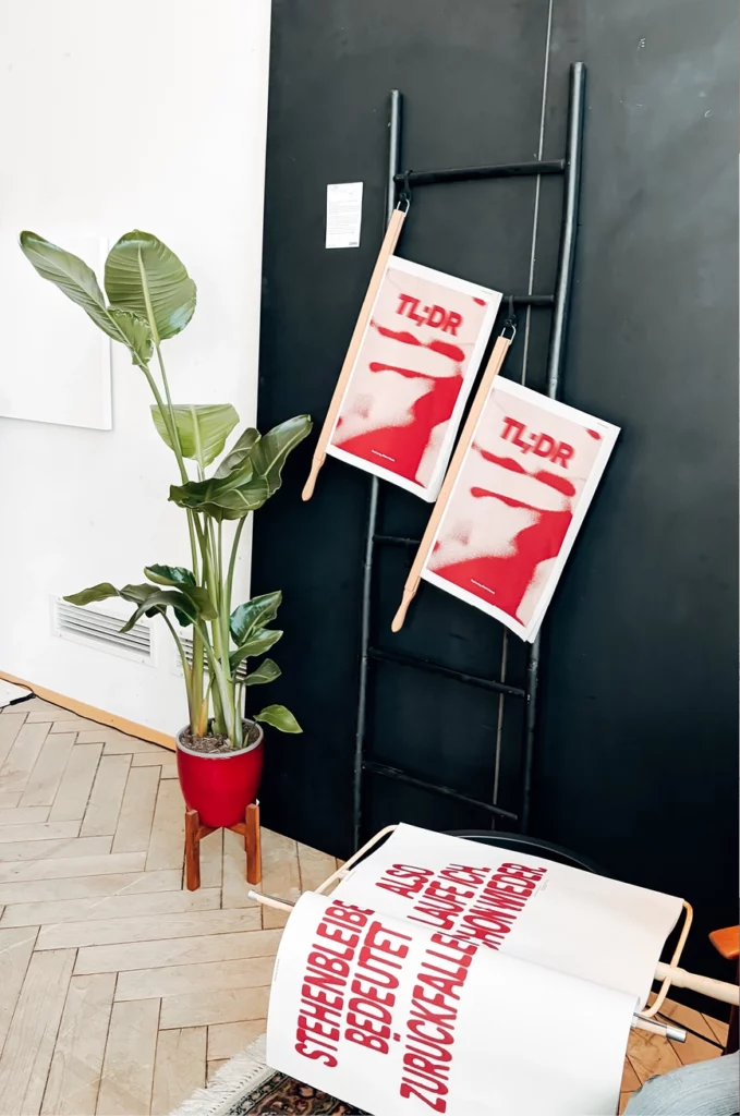 An einer schwarzen Wand lehnt eine dekorative Leiter, an der zwei rot-weiße TL;DR-Poster hängen. Daneben steht eine grüne Blattpflanze in einem Topf, und über einem Stuhl ist ein weißes Tuch mit rotem deutschen Text drapiert.