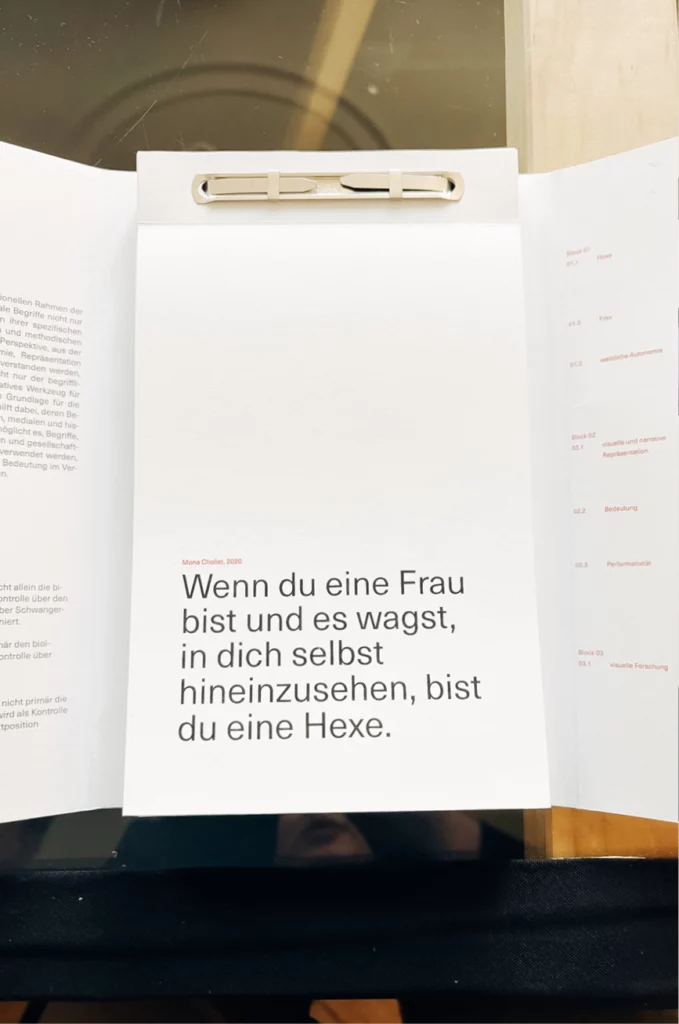Ein Notizblock mit einem deutschen Text, der lautet: „Wenn du eine Frau bist und es wagst, in dich selbst hineinzusehen, bist du eine Hexe.“ Die Seiten werden oben von einer Metallklammer zusammengehalten.