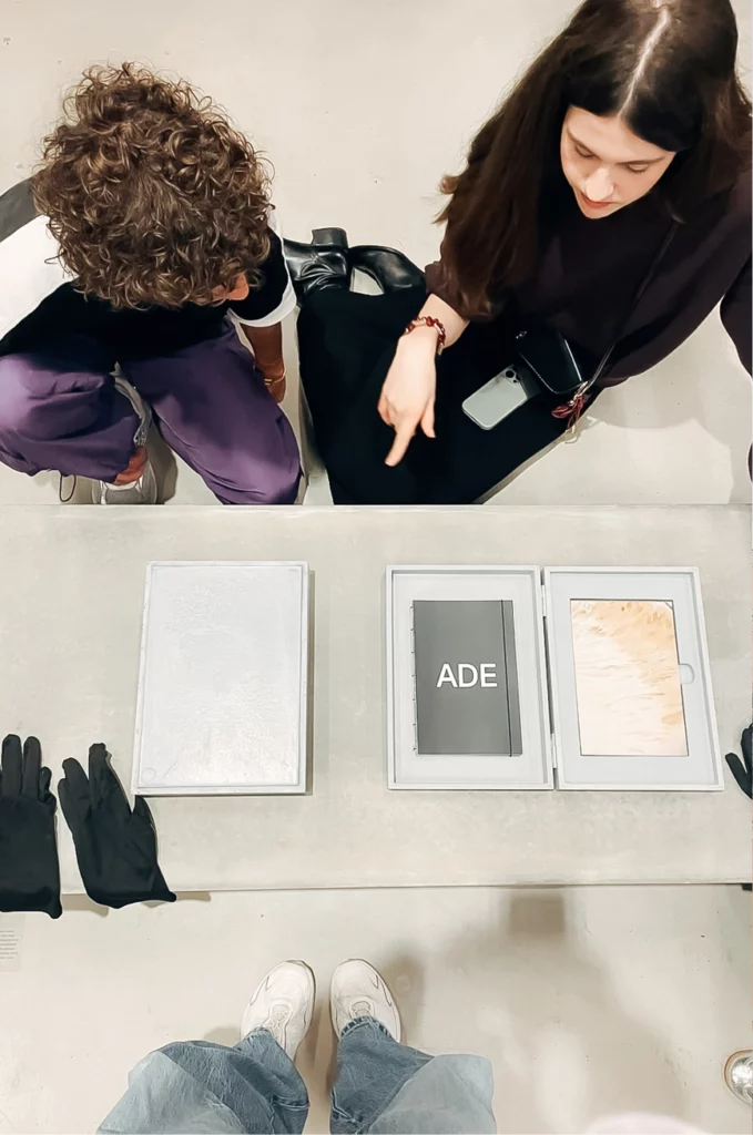 Zwei Personen blicken auf Vitrinen hinab, in denen Bücher liegen. Ein Buch ist auf einer Seite aufgeschlagen, auf der „ADE“ zu lesen ist. Auf der grauen Tischoberfläche sind schwarze Handschuhe und die Spiegelungen ihrer Schuhe zu sehen.