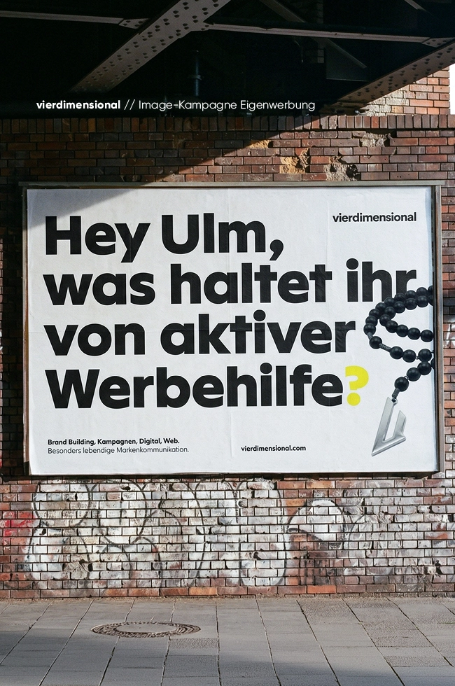 Plakat an einer Backsteinmauer mit der Aufschrift Hey Ulm, was haltet ihr von aktiver Werbehilfe? sowie dem vierdimensional-Logo und einer herunterhängenden Perlenkette.